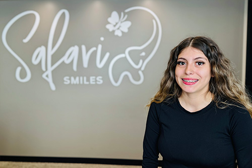 The image shows a woman standing in front of a sign with the word Salavar and a logo that appears to be related to dental care or orthodontics. She is smiling at the camera, wearing a black top, and has long hair.