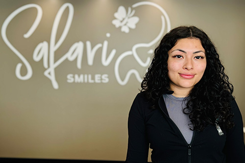 A woman with long brown hair, wearing a black top with white text, stands in front of a sign that reads Salavador Smiles .