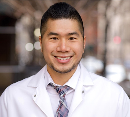 The image is a photograph of a smiling man wearing glasses, a white lab coat over a plaid shirt, and a stethoscope around his neck. He has short dark hair and appears to be standing outdoors with urban architecture in the background.