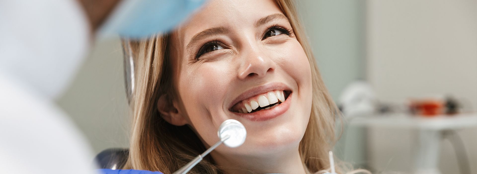 The image shows a woman smiling at the camera while seated in a dental chair, with a dentist visible behind her.